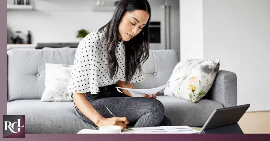A girl is sitting on the couch, writing on a piece of paper in front of her laptop.
