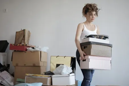 A girl with curly hair tied up, carrying boxes and items, with many stacked boxes on the floor.