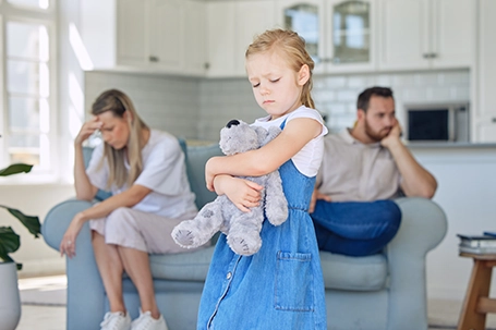 A young girl with a solemn expression stands in the foreground, holding a grey teddy bear close to her chest, while her parents are seated separately on a couch, both looking upset.