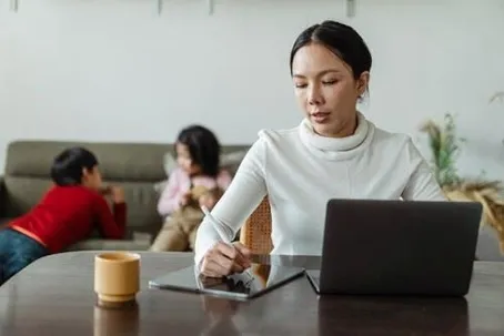 A woman is writing in front of her laptop, with two kids in the background.