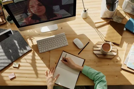 A woman watching her computer while writing at a messy table with a cup of coffee.