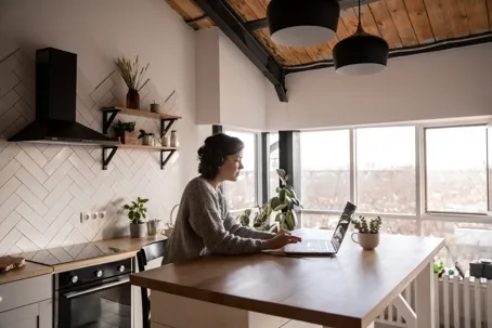 An image of a woman in the kitchen using her laptop placed on the table.