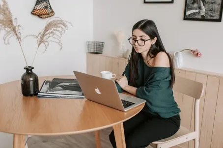 A woman sitting on a chair, wearing eyeglasses, and watching her laptop on the table.