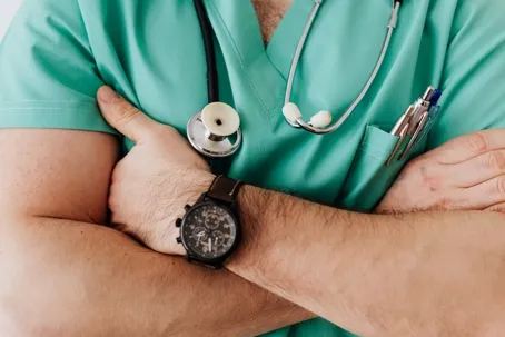 A doctor wearing green scrubs with a stethoscope around his neck and arms crossed.