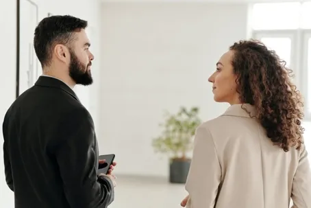 A man and a woman dressed in business attire talking to each other in a bright office hallway.