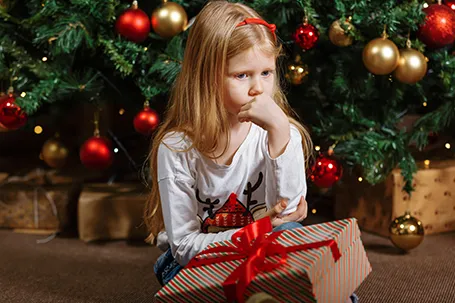 A young girl with long blonde hair sitting in front of a decorated Christmas tree, she has a somewhat sad or pensive expression.