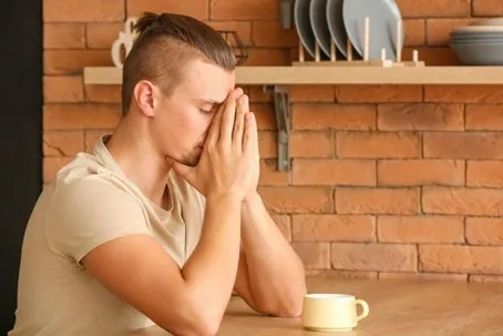 A man sitting at a table with his hands covering his face, looking stressed or upset, with a cup in front of him.