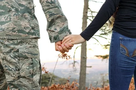 An image of two people holding hands while facing away from the camera — the boy is wearing a military uniform, and the girl is wearing civilian clothes.