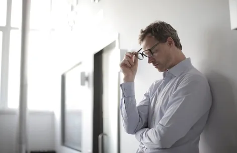 A man wearing eyeglasses and a light-colored shirt leaning against a wall, holding his glasses and looking down with a thoughtful or stressed expression.