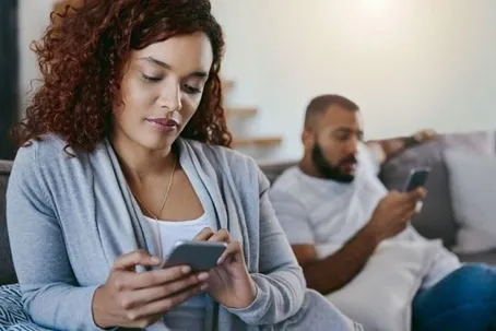 A man and a woman sitting separately on a couch while texting on their phones.