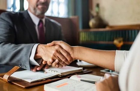 An image of a man wearing a suit and necktie shaking hands with a woman over notes and other documents on the table.