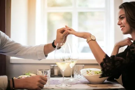 A girl and a boy are eating a meal, while the boy puts a ring on the girl’s hand.