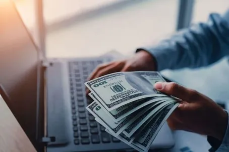 A man holding a lot of money while typing on his computer.