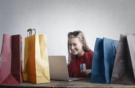 A girl is smiling in front of her laptop, with colorful paper bags on the table.