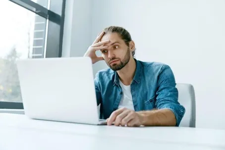 A man is sitting in front of his laptop with his hand on his head.