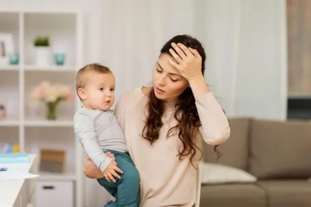 woman is carrying a baby while holding her head, as if she’s not feeling well.