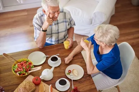 A sweet elderly couple is drinking juice at a table with some food and coffee.