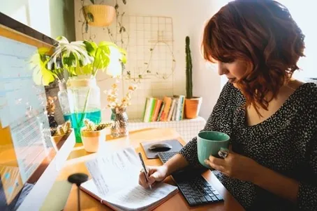 A girl is writing in her notebook while holding a cup in front of the computer.