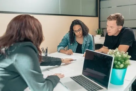 Three people discussing some documents with a laptop and a pot on the table.