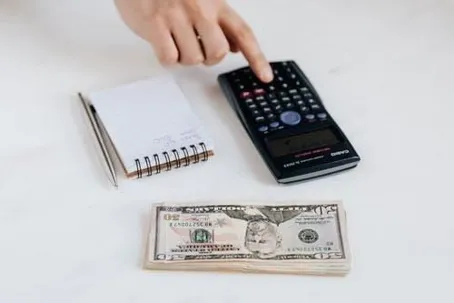 A hand typing on a calculator, with money and a notebook on the table.