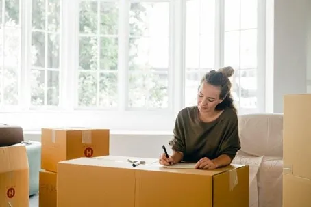 A woman writing on top of a box, surrounded by three more boxes.