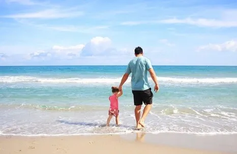 A father and his baby girl walking on the sand by the sea.