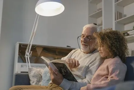 The grandfather is reading a storybook to his grandchild while wearing eyeglasses.