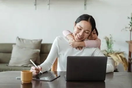 A mother is working on her laptop and writing while her daughter hugs her from behind.