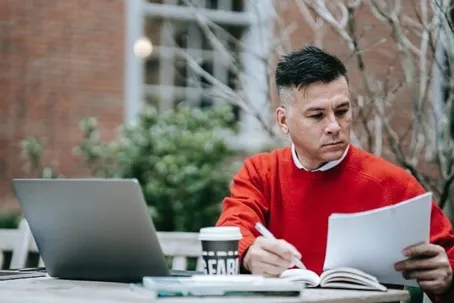 A man reading a document and writing, with a cup of coffee and a laptop on the table.