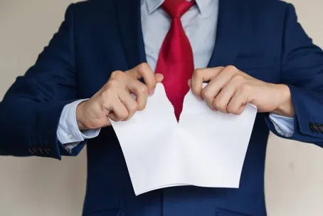 A man wearing a blue suit and a red necktie tearing a piece of paper against a white background.
