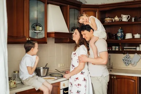 An image of a happy family preparing a meal in the kitchen while the father carries a little one on his shoulders.