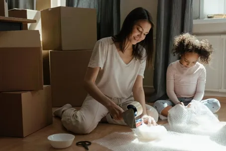 A mother and her daughter sitting on the floor packing items with bubble wrap and boxes around them.