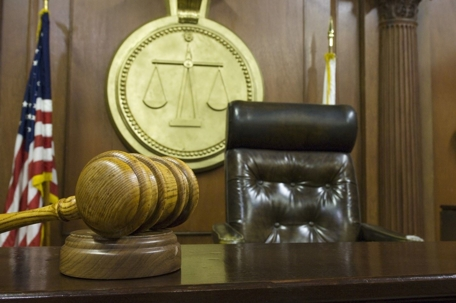 A wooden judge’s gavel on a desk in a courtroom, with an empty judge’s chair and a large justice scale emblem in the background.