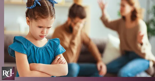 An image of a little girl frowning and looking down, while in the blurry background, her parents are arguing.