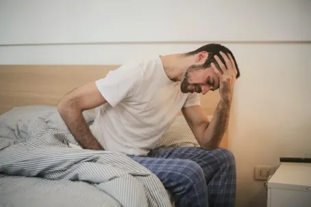 An image of a man who has just woken up, sitting on the edge of the bed, looking stressed while holding his head and facing down.