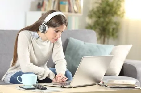 A woman sitting on a couch wearing headphones and watching her personal computer, with a pillow beside her and a cup, cellphones, and notes on the table.
