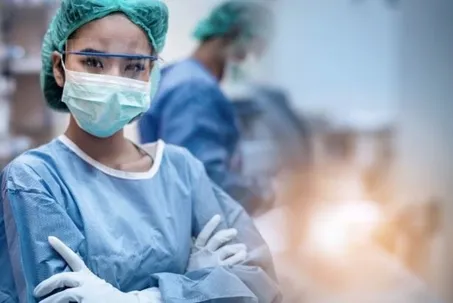 A woman wearing a light blue suit, gloves, face mask, face shield, and hair cap, standing with her arms crossed.