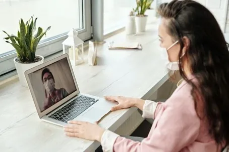 A girl wearing a mask in front of her laptop, making a video call with another person who is also wearing a mask.