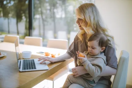An image of a blonde mother carrying her baby while typing on her laptop, with the baby holding and eating an orange.