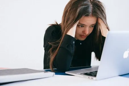A girl has a worried expression as she faces her laptop, with her hands in her hair.