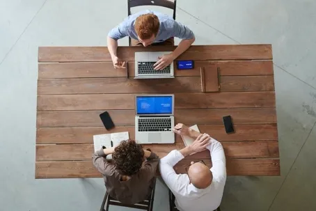 Three people sitting at a wooden table having a meeting while working on their laptops.