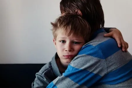 A young boy with a sad face, hugging his father.