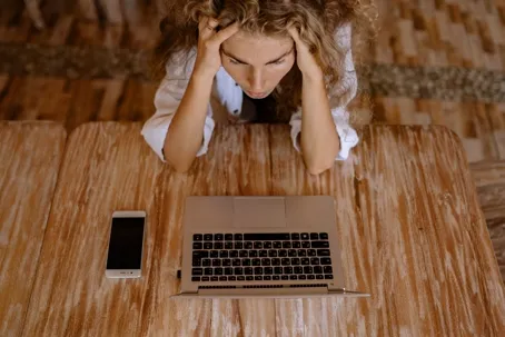 A woman feeling irritable and stressed, sitting at a table with her hand pulling her hair, in front of a laptop with a cellphone placed behind it.