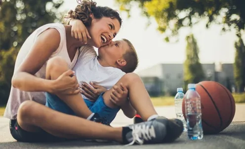 An image of a mother and her son playfully bonding, with the son kissing his mother, and two bottles of water and a ball nearby.