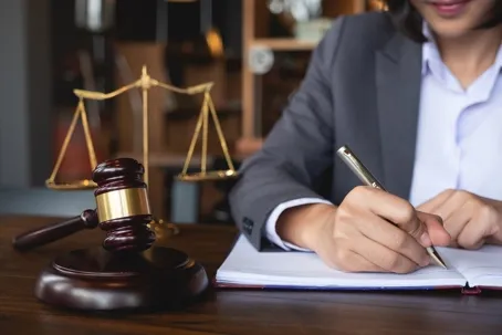 An image of a woman wearing a suit and writing, with a gavel in front of her and a justice scale beside her.