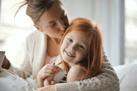 A mother kisses her blonde-haired daughter who is smiling.