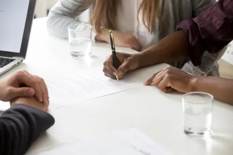 Two people are sitting beside each other and signing a document, with a lawyer seated in front of them.