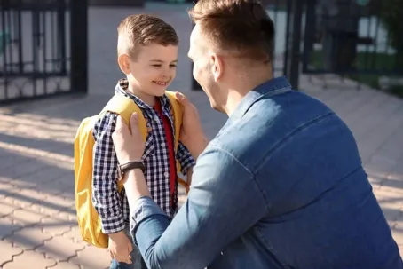 An image of a father and son both smiling, as the father adjusts his son’s backpack before going to school.