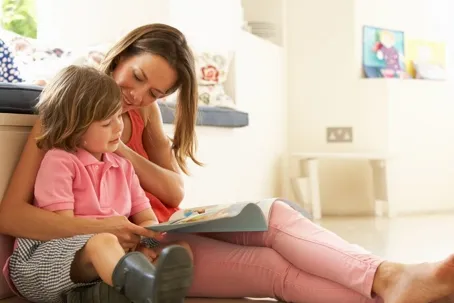 An image of a mother and daughter reading a book while sitting on a couch.