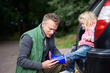 A man putting shoes on his little girl who is sitting in the back of the car.